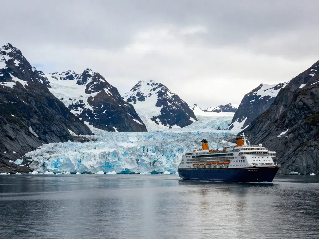 A cruise ship passing a blue glacier beneath snow-capped mountains in an Alaskan fjord